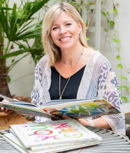 Portrait shot of Alex outdoors. Alex is a middle aged woman with blond hair and a big smile, wearing a black top and patterned shrug, sitting outside with a pile of books