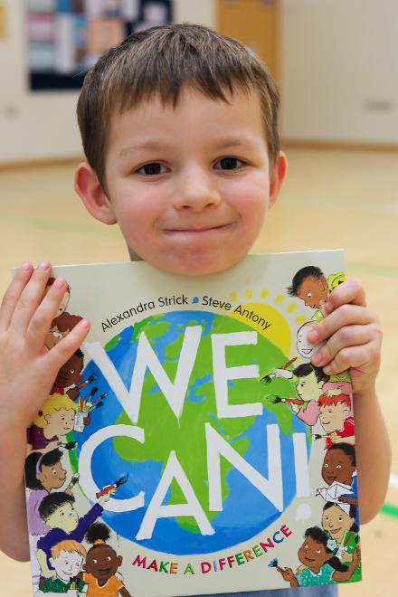 A close-up of a small boy with dark hair holding a copy of WE CAN under his chin, slightly smiling